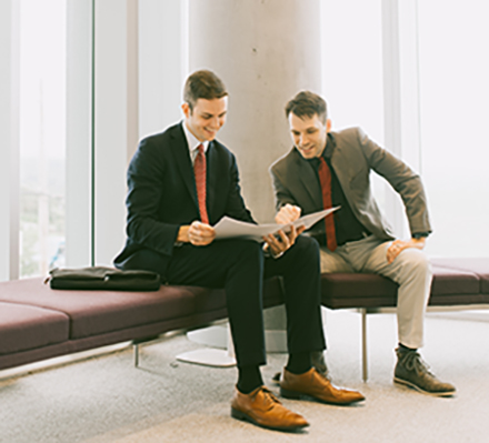 two students on a bench wearing suits and pointing to an academic paper and smiling